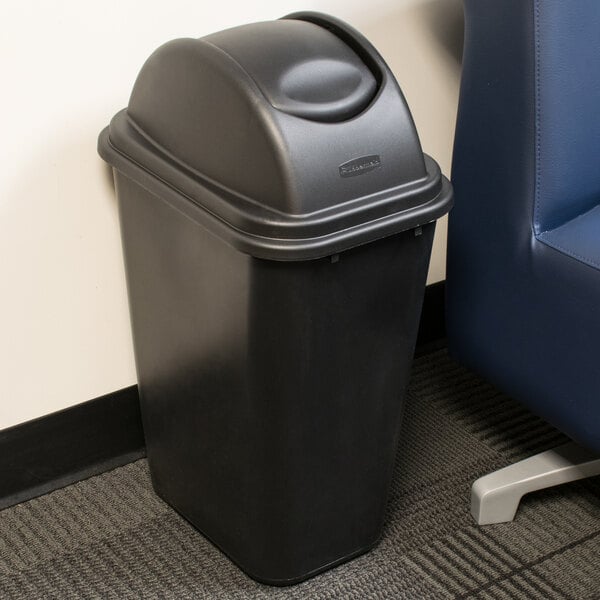 A black Rubbermaid Untouchable trash can with a swing lid next to a blue chair.