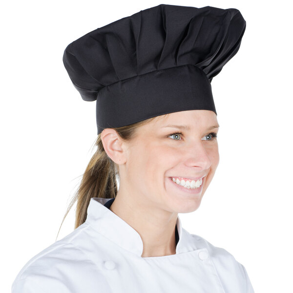 A woman in a black Chef Revival chef's hat smiling while standing at a counter in a professional kitchen.