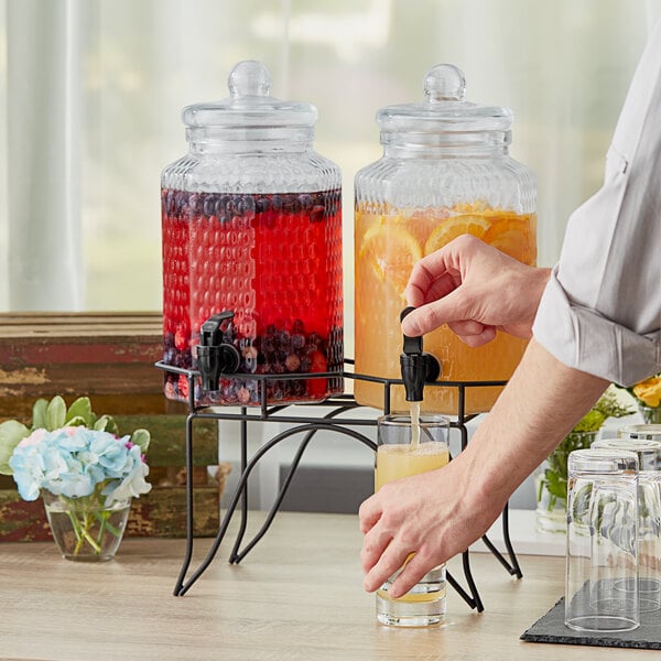 A person pouring a drink from an Acopa double glass beverage dispenser with metal stand.