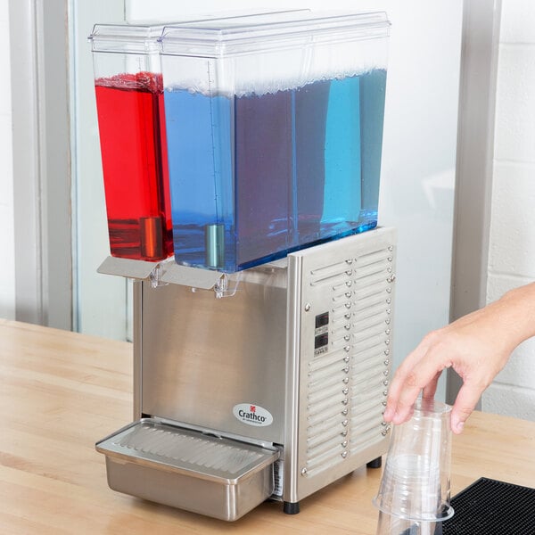 A hand pouring liquid into a Crathco Mini-Twin stainless steel cold beverage dispenser.