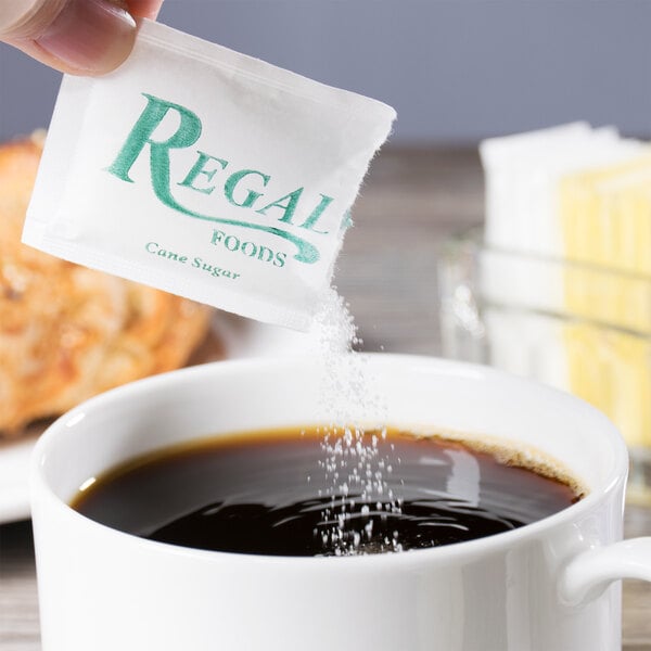 A hand holding a Regal cane sugar packet over a cup of coffee with brown liquid and sugar.