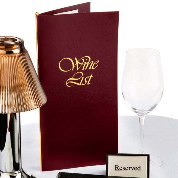 A burgundy Menu Solutions wine list cover on a table with a wine glass and a wine list.