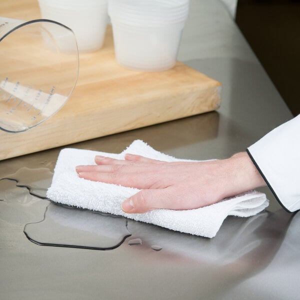 A person using a white Chef Revival bar towel to wipe a glass on a wood counter.