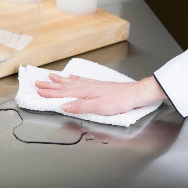 A hand with a Chef Revival white terry bar towel on a counter wiping a surface.