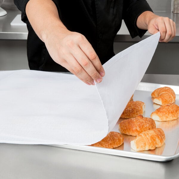 A hand using a Choice white flour sack towel to cover a tray of croissants.