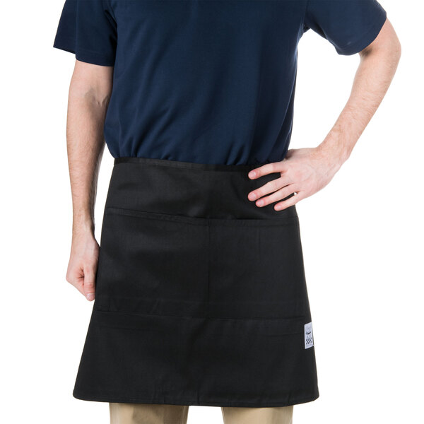A man wearing a black Chef Revival bistro apron with 2 pockets standing in a professional kitchen.