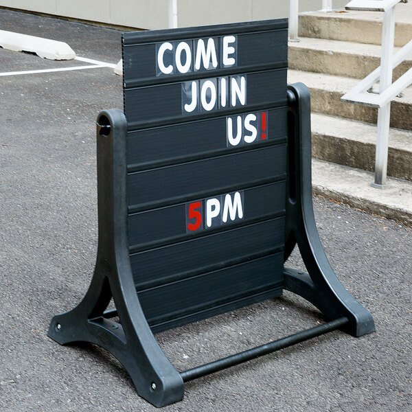 A black Aarco Rocker letterboard on a sidewalk with white letters reading "Come Join Us 5 pm"