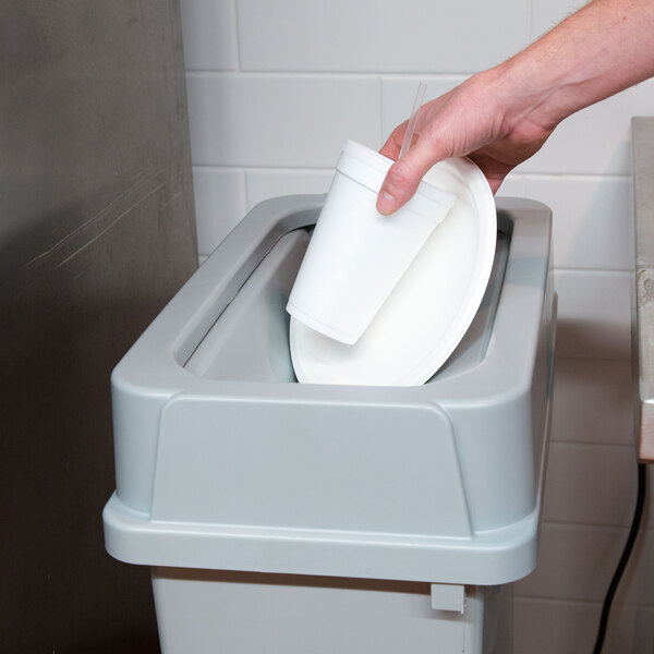 A hand holding a white cup dropping it into a gray rectangular trash can.
