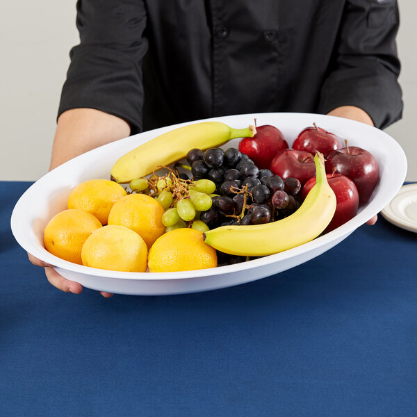 A Fineline white plastic deep oval bowl filled with bananas, oranges, and grapes on a table.