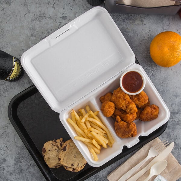 A white styrofoam takeout container with 2 compartments holding chicken and fries with a bottle of sauce.