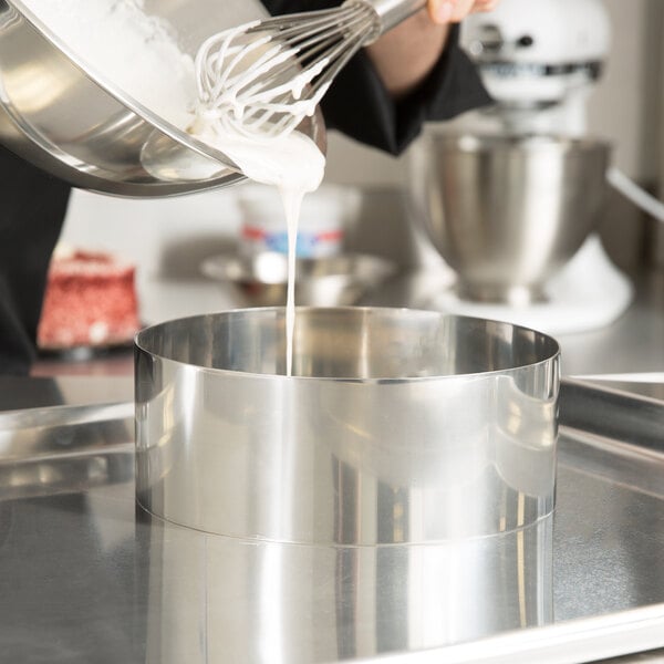 A person using an American Metalcraft stainless steel round cake ring to mix white liquid in a bowl with a whisk.