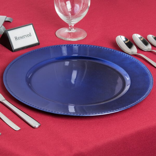 A blue Charge It by Jay beaded plastic charger plate with silverware on a red table.