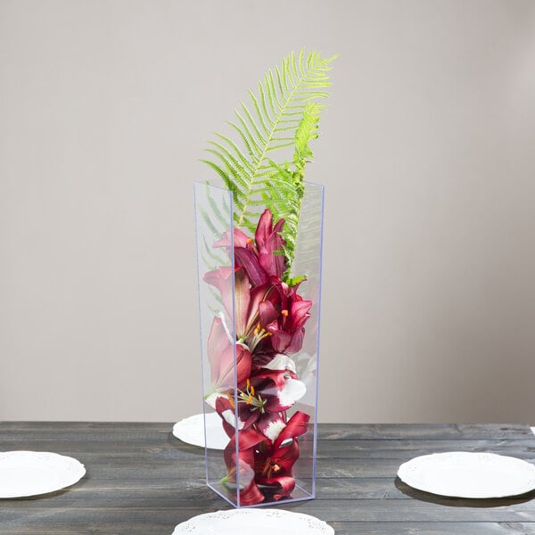 A clear square acrylic vase with red flowers and a green leaf on a table.