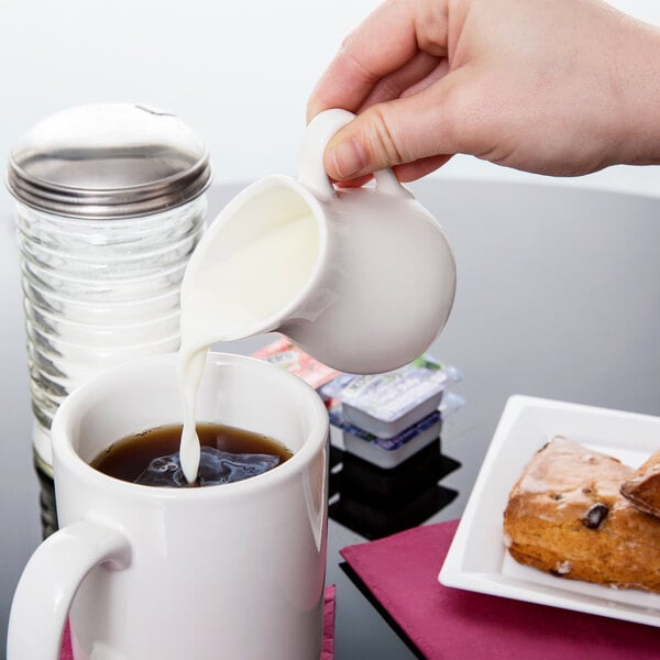 A hand pouring white liquid from an American Metalcraft porcelain bell creamer into a mug of coffee.