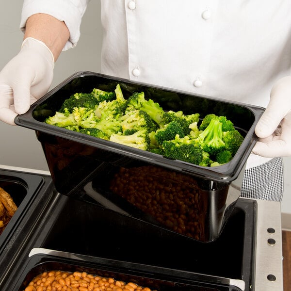 A person in a white coat holding a Cambro black plastic food container full of broccoli.