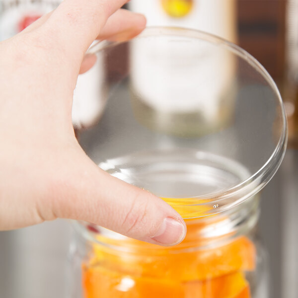 A person's hand holding an American Metalcraft Mason Jar with an orange drink and a clear plastic lid.