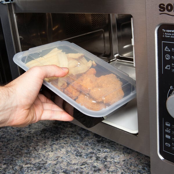 A hand holding a Pactiv plastic container of chicken and french fries in a microwave.