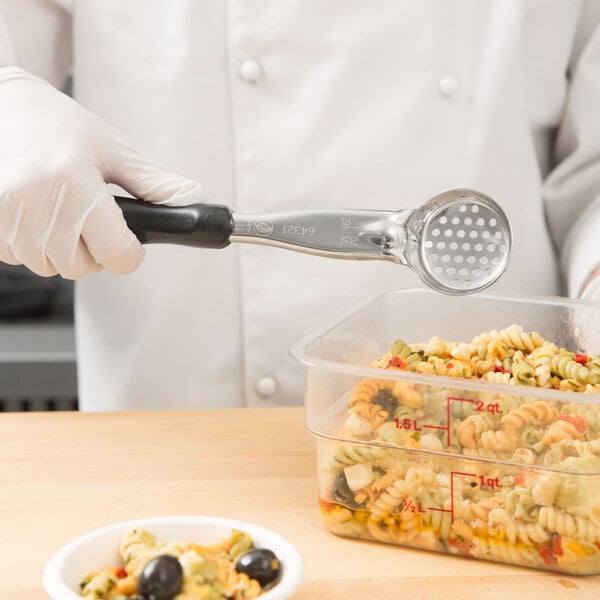 A gloved hand uses a Vollrath black perforated round Spoodle to serve pasta from a plastic container.