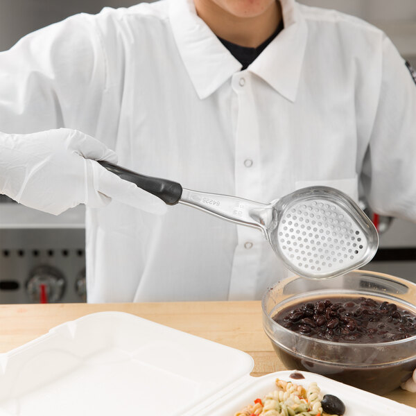 A person in a white coat using a Vollrath black perforated oval Spoodle to serve food into a bowl.
