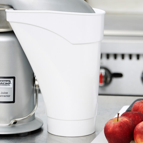 A white Waring pulp container with a black lid next to a juice extractor filled with apples.