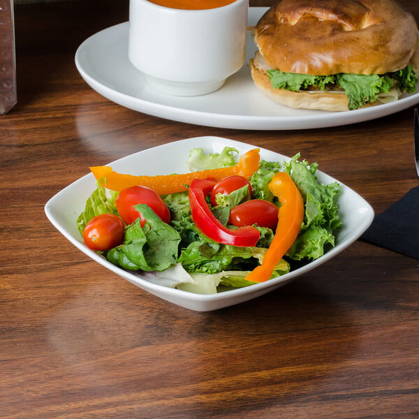 A CAC square bowl of salad with tomatoes and peppers next to a sandwich on a table.