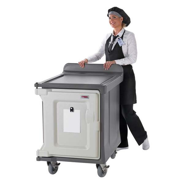 A woman in a chef hat and apron pushing a large granite gray Cambro meal delivery cart.