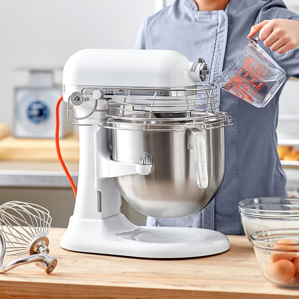 A woman pouring eggs from a glass bowl into a white KitchenAid countertop mixer.