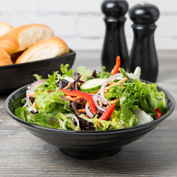 A black Milano melamine bowl filled with salad on a table with bread and wine.
