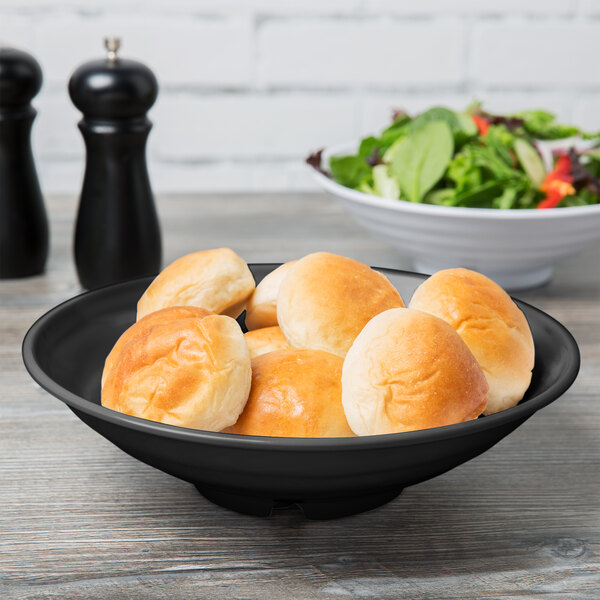 A black Milano melamine bowl filled with bread rolls on a table with a bowl of salad.