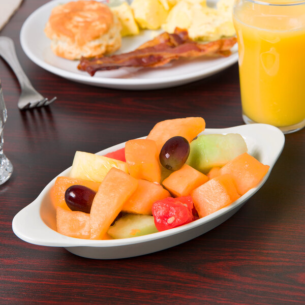 An ivory oval bowl filled with food on a table with a plate of fruit and a glass of orange juice.
