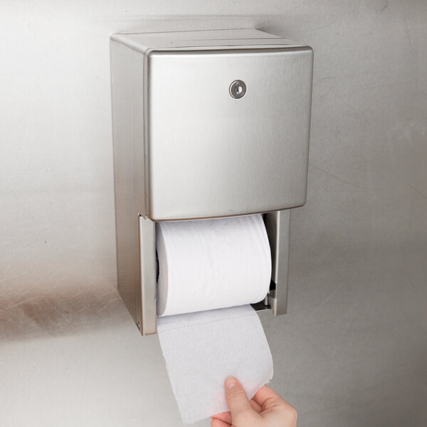 A hand pulling a roll of toilet paper out of a Bobrick multi-roll toilet paper dispenser on a wall.