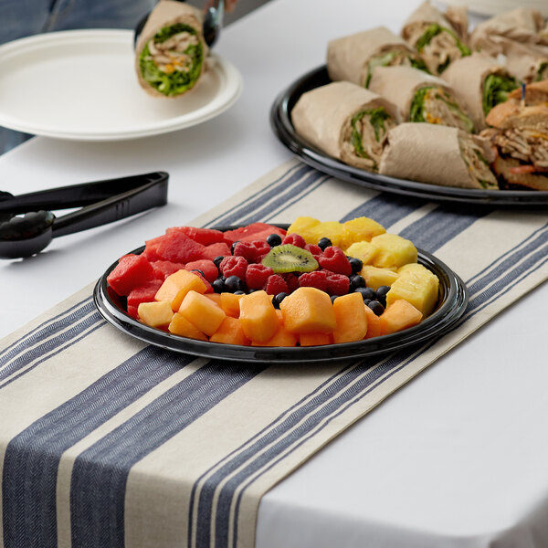 A Visions black plastic catering tray filled with fruit and sandwiches on a table with a striped tablecloth.