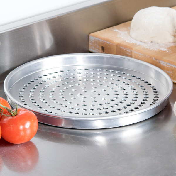 An American Metalcraft heavy weight aluminum super perforated pizza pan on a white surface with a ball of dough and tomatoes.