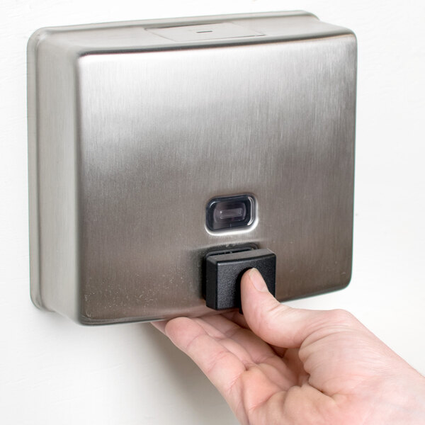 A hand pressing a button on a silver wall mounted Bobrick soap dispenser.