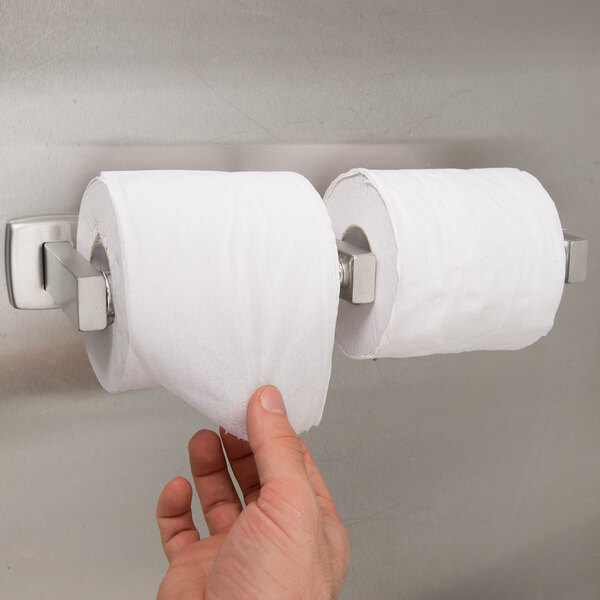A hand reaching for a roll of toilet paper in a metal toilet paper holder.