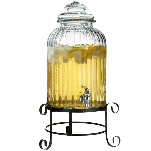 A Stylesetter glass beverage dispenser with metal stand filled with a yellow drink and ice.