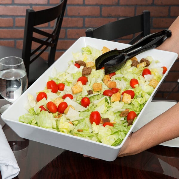 A person holding a white tray of salad with a pair of tongs over a white rectangular bowl.