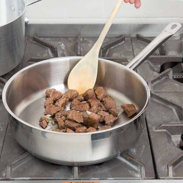 A person stirring meat in a Vollrath saute pan with a wooden spoon.