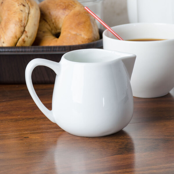A CAC white porcelain creamer pitcher on a wood surface next to a white cup of coffee and bagels.