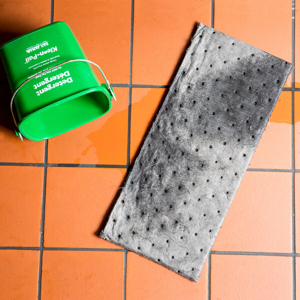 A green bucket with white text and a grey absorbent pad on a tile floor.