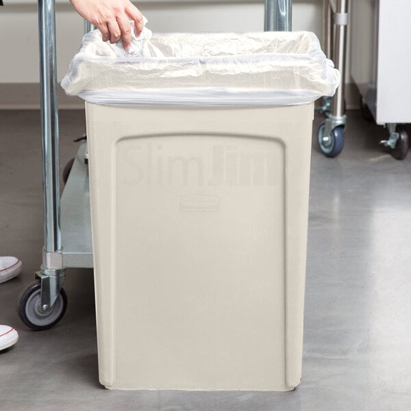A woman putting a plastic bag in a Rubbermaid Slim Jim Beige rectangular trash can.