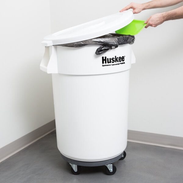 A person putting a green trash bag into a white Continental Huskee trash can with a green lid.