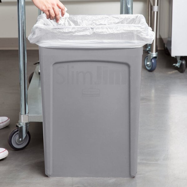 A person using a clear plastic bag to line a Rubbermaid Slim Jim trash can.