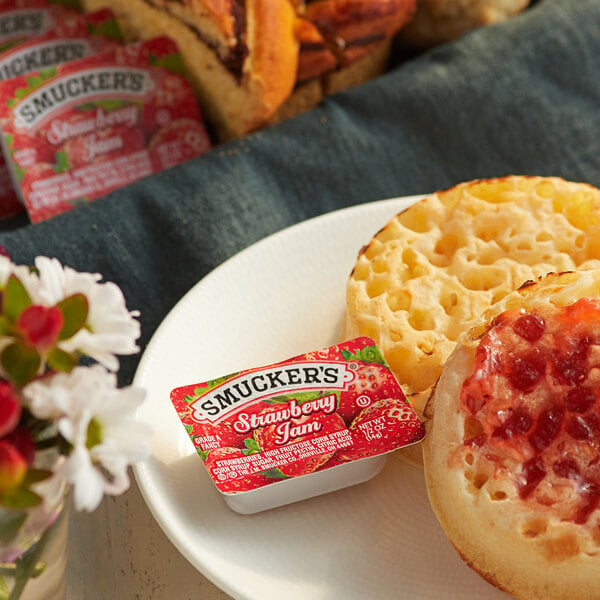 A plate with a bagel and a Smucker's strawberry jam cup on a table of food.