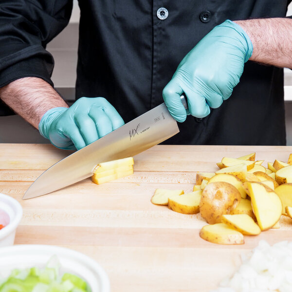 A person wearing blue gloves uses a Mercer Culinary MX3 Japanese Gyuto knife to cut potatoes on a table.