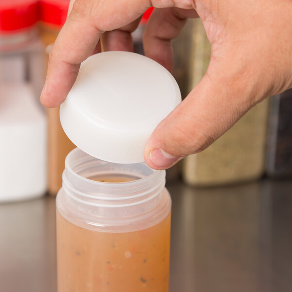 A hand holding a plastic container with a white Tablecraft cap pouring liquid into another container.