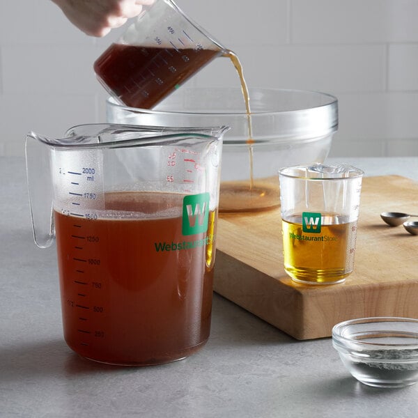 A hand pouring brown liquid into a WebstaurantStore clear plastic measuring cup.