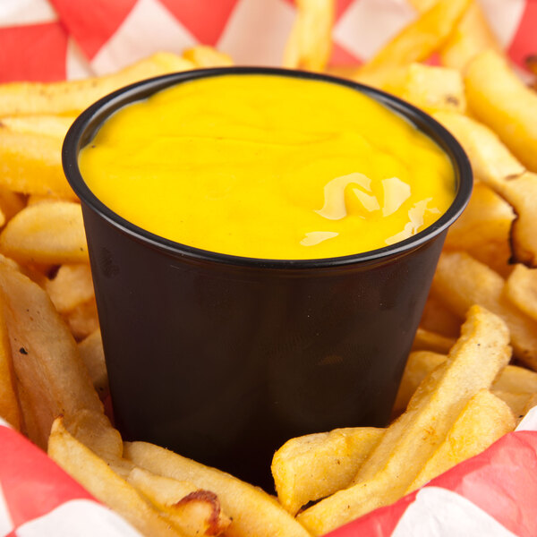 A black Solo portion cup filled with yellow sauce on a basket of french fries.