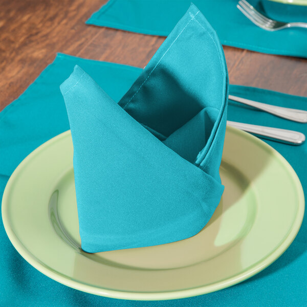 A folded teal Intedge cloth napkin on a plate with silverware.
