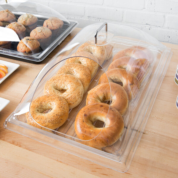 A clear polycarbonate tray with a bagel with sesame seeds on it and other bagels on a table in a bakery display.
